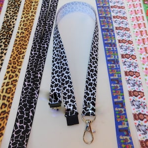 May include: A set of colorful patterned lanyard straps with a black and white cow print lanyard strap with a silver swivel clip. The straps are made of fabric and have a variety of patterns, including animal prints, geometric patterns, and cartoon characters.