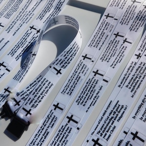 Könnte beinhalten: Weiße Satinband-Lanyards mit schwarzen Kreuzen und inspirierenden Bibelversen darauf gedruckt. Die Lanyards sind in einem Stapel angeordnet, einige davon sind umgeklappt.