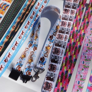 May include: A collection of colorful lanyards with various patterns. Designs include roosters, horses, cowboys, and unicorns. Some lanyards have text, and all have a clasp and a black plastic clip.