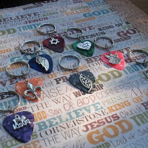May include: A collection of guitar pick keychains with various charms. The picks are in assorted colors, and the charms include a Star of David, an angel, a fleur-de-lis, and the words "I Love God" and "I Jesus".