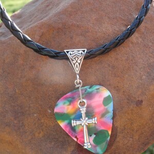 May include: A colorful guitar pick pendant necklace. The pendant features a silver cross and a multi-colored pick with pink, green, blue, and yellow hues. The necklace has a black braided leather cord.