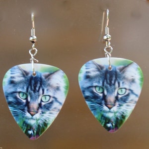 May include: A pair of earrings featuring a cat's face printed on a teardrop-shaped pendant. The cat has green eyes and gray and white fur. The earrings have silver-colored hooks.