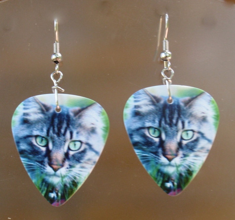 May include: A pair of earrings featuring a cat's face printed on a teardrop-shaped pendant. The cat has green eyes and gray and white fur. The earrings have silver-colored hooks.