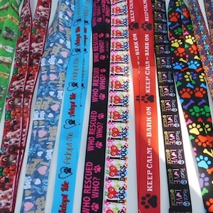 May include: A collection of dog-themed lanyards with various patterns and text. Designs include dog breeds, paw prints, hearts, and phrases like "Adopt Me," "Who Rescued Who?" and "Keep Calm and Bark On." Colors include green, red, blue, and black.