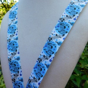 May include: A blue and white lanyard featuring a musical note pattern. The lanyard has a repeating design of musical notes and treble clefs in black against a light blue background with blue polka dots. The lanyard is displayed on a white mannequin.