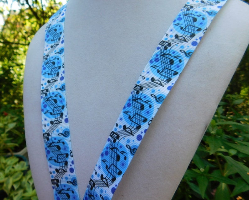 May include: A blue and white lanyard featuring a musical note pattern. The lanyard has a repeating design of musical notes and treble clefs in black against a light blue background with blue polka dots. The lanyard is displayed on a white mannequin.