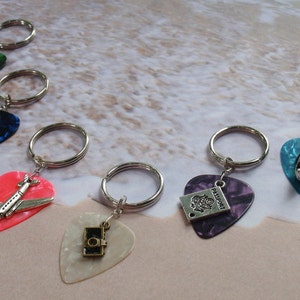 May include: Keychains featuring guitar pick shapes in various colors, each with a silver charm. Charms include a camera, airplane, Eiffel Tower, boat, and passport. The keychains have silver key rings and are set against a neutral background.