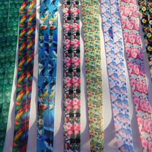 May include: A collection of colorful lanyards with various patterns. Designs include peacock feathers, tropical birds, hibiscus flowers, and fish. The lanyards are made of a smooth, shiny material and are displayed side by side.