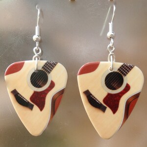 Puede incluir: Un par de pendientes de p&uacute;a de guitarra. Cada pendiente presenta una p&uacute;a de color crema con un dise&ntilde;o de guitarra ac&uacute;stica impresa, que incluye una boca roja y detalles del diapas&oacute;n. Los pendientes cuelgan de alambres de gancho plateados.