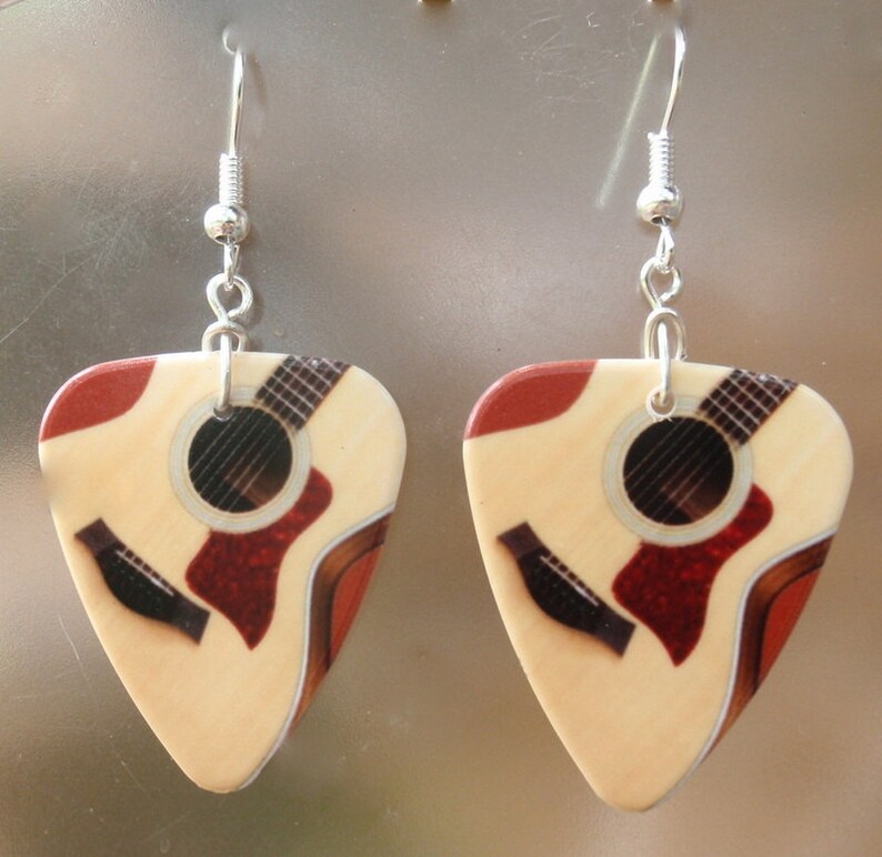 Puede incluir: Un par de pendientes de p&uacute;a de guitarra. Cada pendiente presenta una p&uacute;a de color crema con un dise&ntilde;o de guitarra ac&uacute;stica impresa, que incluye una boca roja y detalles del diapas&oacute;n. Los pendientes cuelgan de alambres de gancho plateados.