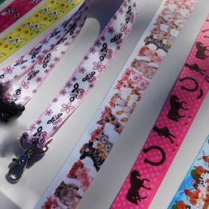 May include: A collection of colorful lanyards with various patterns. Designs include cows, flowers, horseshoes, and horses. The lanyards are made of ribbon and have a metal clasp and black plastic clip.