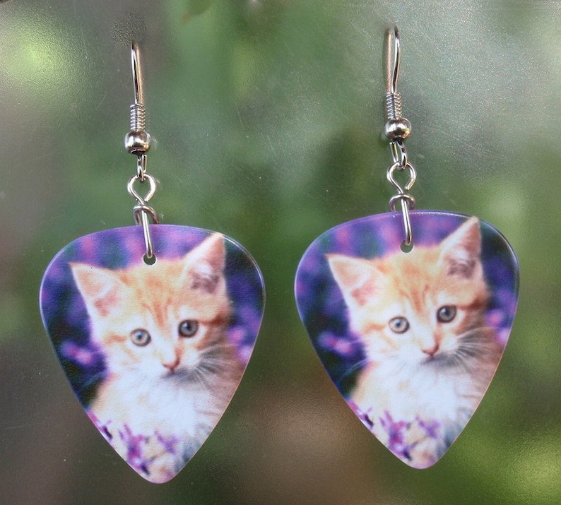 May include: A pair of earrings featuring a kitten design. The earrings are shaped like guitar picks, with a light orange kitten on a purple background. The earrings have silver-colored hooks.