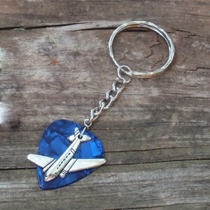 May include: A blue guitar pick keychain with a silver airplane charm. The keychain has a silver chain and a silver key ring. The guitar pick has a textured, shiny surface.