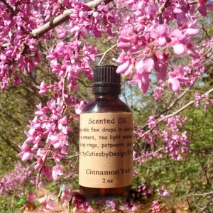 May include: A brown glass bottle of "Cinnamon Yum" scented oil with a black cap. The label reads "Scented Oil" and "2 oz." The background features pink blossoms and green foliage.