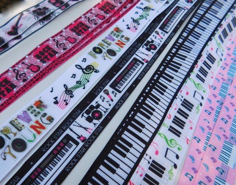 May include: A collection of musical-themed ribbons in various designs. Ribbons feature musical notes, piano keys, guitars, and the text "ROCK STAR" and "FOREVER YOUNG". Colors include pink, black, white, and green. Ideal for crafting or music-related projects.
