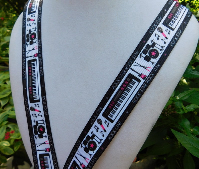 May include: Black and white ribbon with a musical theme. The ribbon features illustrations of musical instruments like keyboards, drums, and microphones, along with musical notes. The words "ROCK STAR" are printed repeatedly along the edges of the ribbon.