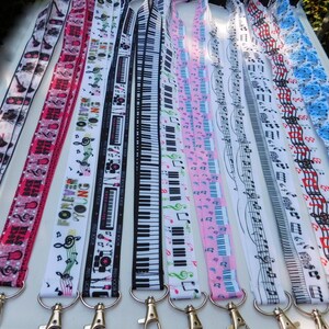 May include: A collection of lanyards with various music-themed designs. The lanyards feature patterns of guitars, musical notes, piano keys, and the text "Forever Young" in a variety of colors, including pink, white, and blue. Each lanyard has a silver-colored metal clasp.