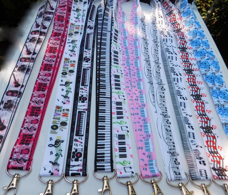 May include: A collection of lanyards with various music-themed designs. The lanyards feature patterns of guitars, musical notes, piano keys, and the text "Forever Young" in a variety of colors, including pink, white, and blue. Each lanyard has a silver-colored metal clasp.