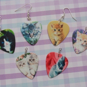 May include: Six pairs of cat-themed earrings. Each earring is a guitar pick shape with a different cat portrait. The cats are various colors, including black and white, orange, and gray. The earrings have silver-colored hooks.