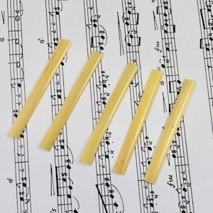 Puede incluir: Cuatro cañas de clarinete amarillas dispuestas en diagonal sobre una hoja de música. Las cañas están hechas de caña natural y se utilizan para producir sonido en un clarinete.