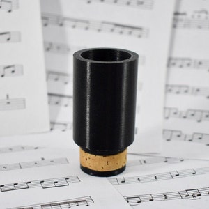 Könnte beinhalten: Schwarzes Klarinettenmundstück mit Korkbasis. Das Mundstück liegt auf einem Blatt Musik.