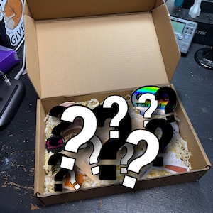 May include: A cardboard box filled with various stickers and other items, including a holographic sticker. The box is open, revealing the contents, which are nestled in paper filler. The stickers have various colors and designs.