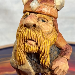May include: A hand-carved wooden figurine of a Viking warrior with a long, flowing beard and a horned helmet. The figurine is holding a shield and an axe. The figurine is painted in shades of brown, yellow, and red.