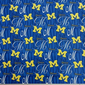 May include: Blue fabric with a repeating pattern of the letter "M" in yellow, along with the words "GO BLUE". The script "M" is in white. The fabric is likely for crafting or apparel.