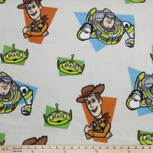 May include: White fabric featuring cartoon characters Woody, Buzz Lightyear, and the Little Green Men from Toy Story. The characters are set against orange, green, and blue geometric shapes. The fabric is ideal for children's crafts.