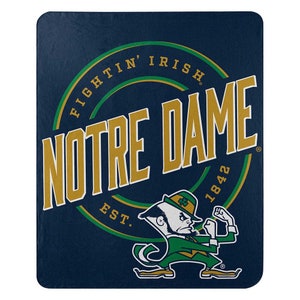 May include: A navy blue fleece blanket with a gold and green Notre Dame Fighting Irish logo. The logo features the words "Fightin' Irish" and "Notre Dame" in a circle with the year "1842" and "Est." below. A cartoon leprechaun is in the bottom right corner.