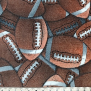 May include: A brown fleece fabric featuring a repeating pattern of American footballs with white laces and grey stripes. A white measuring tape is visible at the bottom, showing measurements from 19 to 31 inches along the edge of the fabric.