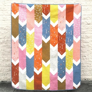 May include: A colorful chevron quilt with a variety of bright colors, including pink, yellow, orange, blue, and brown. The quilt is made up of a series of chevron patterns, each with a different color.
