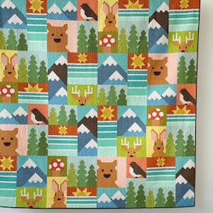 May include: A colorful patchwork quilt featuring woodland creatures and mountain scenes. The design includes bears, rabbits, birds, trees, mushrooms, and suns. The quilt is made of squares in shades of blue, green, orange, and brown.