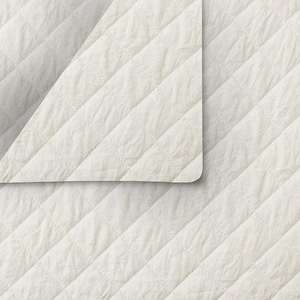 May include: Off-white quilted fabric with a diamond pattern. The fabric is folded over, revealing a rounded corner. The texture appears soft and slightly wrinkled, suggesting a comfortable textile for bedding or upholstery.