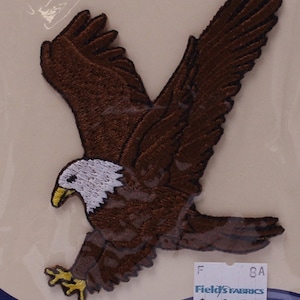 May include: Brown and white embroidered iron-on patch of a bald eagle with wings spread, flying. The patch is packaged on a card with the text "Wrights" and "Appliqué Application".