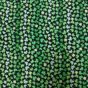 May include: Fabric with a repeating pattern of green shamrocks on a black background. The shamrocks vary in shades of green and some have a pale yellow hue. The text "One Yard" is visible along the bottom edge.