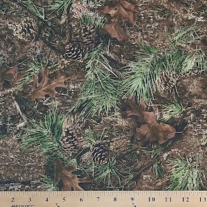 May include: A close-up of a brown and green camouflage fabric with a repeating pattern of pine needles, pine cones, and oak leaves.