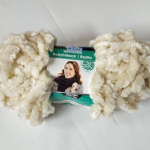 May include: A skein of white yarn with gold glitter threads. The yarn is called "Glimmer" and is labeled as "Scintillant | Brillo".