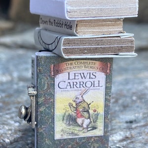 May include: A stack of miniature books with a green cover featuring a white rabbit illustration and the text "The Complete Illustrated Works of Lewis Carroll". The top book has the title "Down the Rabbit Hole" printed on the cover.