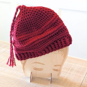 May include: A burgundy crochet hat with a tassel, displayed on a wooden head-shaped stand. The hat has a ribbed brim and a textured, close-knit design. The tassel hangs from the top of the hat.