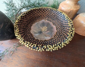 以下が含まれることがあります： 濃い茶色と黒の模様が特徴的な装飾用の織りボウル。縁には金色のビーズがアクセントとして施されています。中央には金色のアクセントが付いた樹脂のデザインがあります。ユニークな手作りの室内装飾品です。