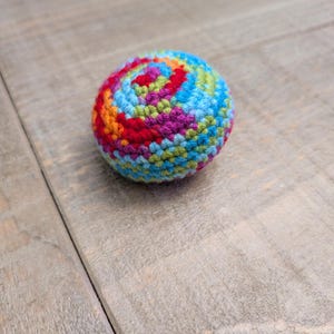 May include: A brightly coloured, crocheted ball with a spiral design. The ball is made from red, orange, yellow, green, blue, and purple yarn. The handmade sphere is on a wooden surface.