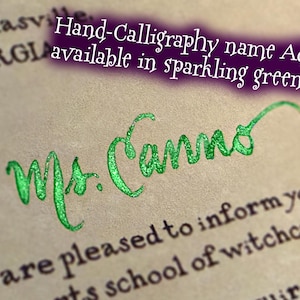 May include: A piece of parchment paper featuring printed black text and the handwritten name Ms. Cannon in sparkling green ink. A purple text box overlay reads Hand-Calligraphy name Add-on available in sparkling green ink!