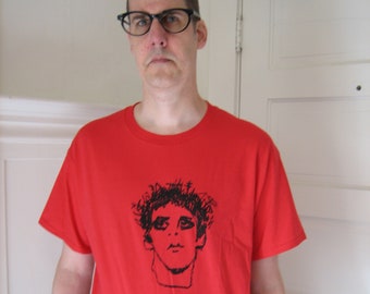 May include: A red t-shirt with a black and white illustration of a person's face on the front.