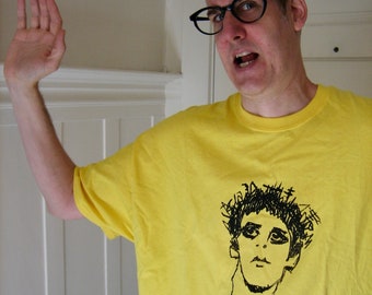 May include: A man wearing a yellow t-shirt with a black and white line drawing of a person's face on the front. He is wearing black framed glasses and has a surprised expression on his face.