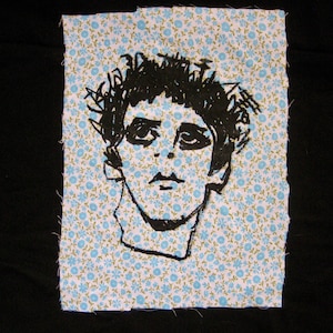 May include: A black and white sketch of a person's face is centered on a small piece of fabric with a light blue floral pattern. The fabric is set against a black background.