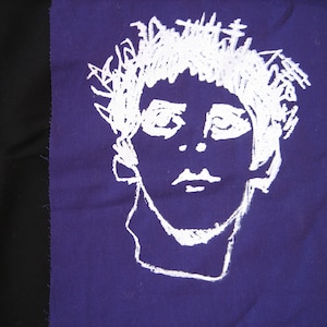 May include: White embroidered portrait of a person with a messy hairstyle on a purple fabric background.