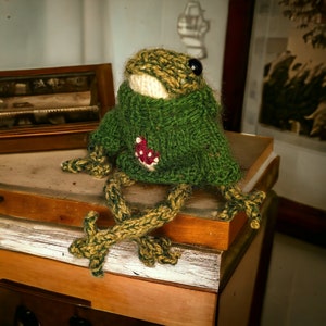 May include: A green knitted frog wearing a green sweater with a red mushroom design. The frog is sitting on a wooden surface.