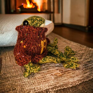 May include: A brown and green knitted frog toy with a black button eye. The frog is sitting on a brown rug.