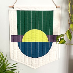 May include: Geometric wall hanging with a cream-colored background and a design featuring a green rectangle, a yellow semi-circle, a purple stripe, and a blue semi-circle. The design is quilted with white stitching and hangs from a wooden dowel.